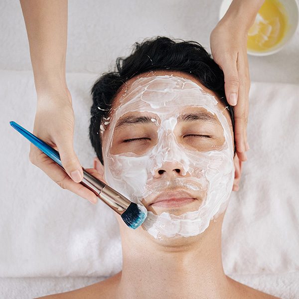 Beautifcian using brush when applying moisturizing mask on face of Asian customer, view from above