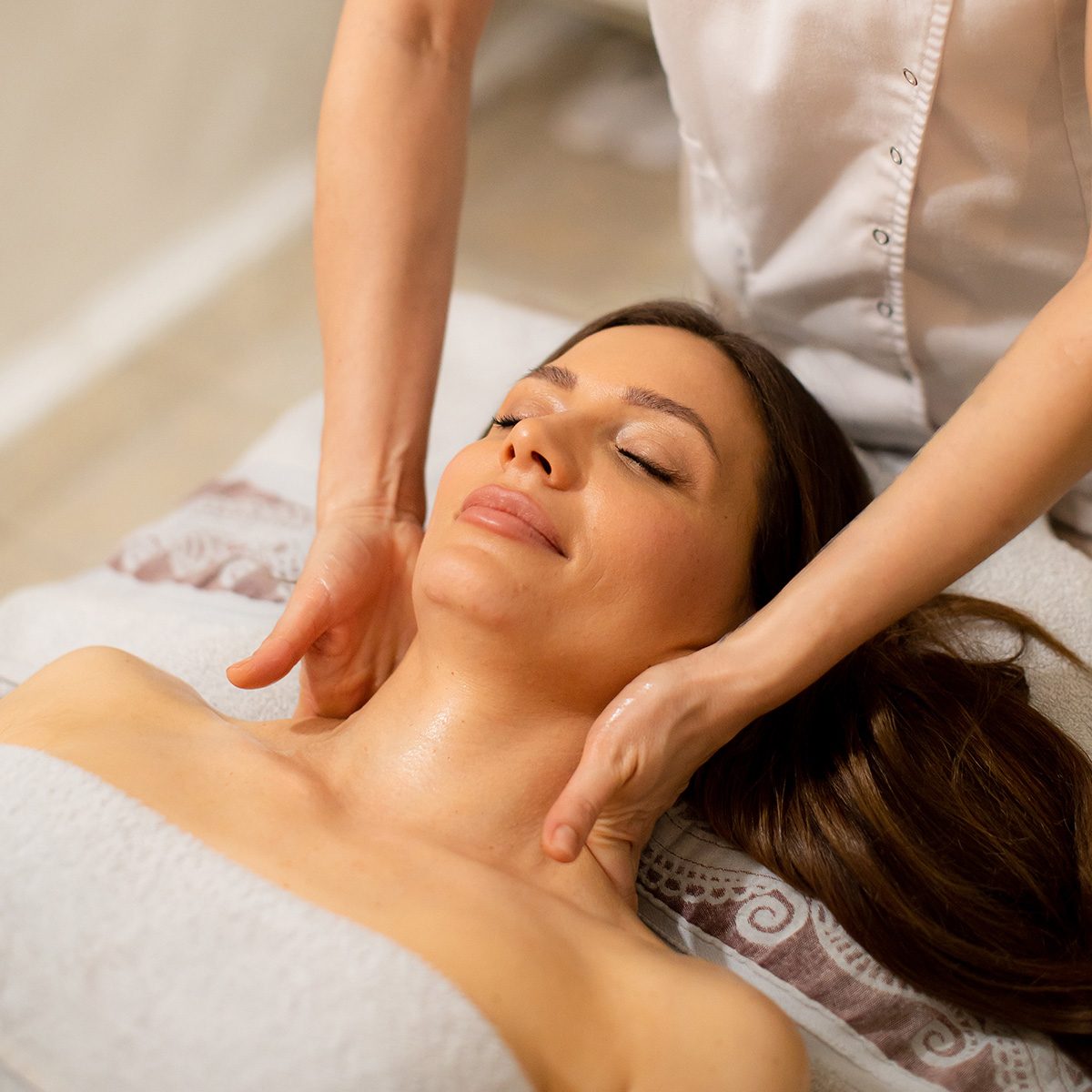 A woman lies on a massage table with a towel around her. A therapist is giving her a neck massage. The room has soft lighting and a relaxing atmosphere.